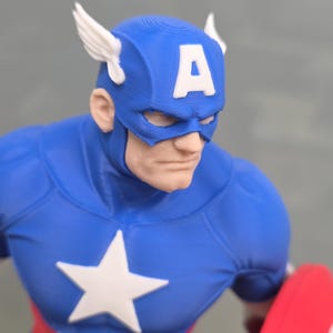 May include: A 3D printed figurine of Captain America featuring a blue mask with white wings and a white letter A on the forehead. The character wears a blue suit with a white star on the chest and red and white stripes at the waist, holding a red shield.