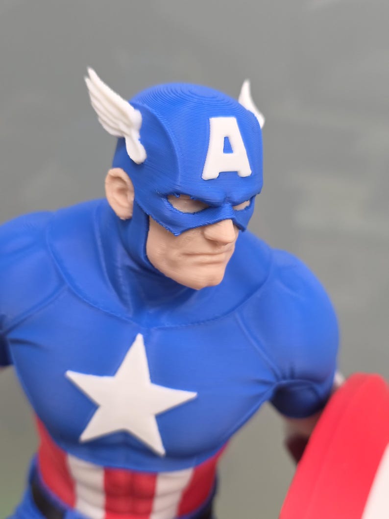 May include: A 3D printed figurine of Captain America featuring a blue mask with white wings and a white letter A on the forehead. The character wears a blue suit with a white star on the chest and red and white stripes at the waist, holding a red shield.