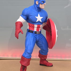 May include: A Captain America action figure wearing a blue suit with a white star on the chest, red and white striped midsection, red gloves, and red boots. The figure wears a blue cowl with a white 'A' and wings, and holds a round red, white, and blue shield.
