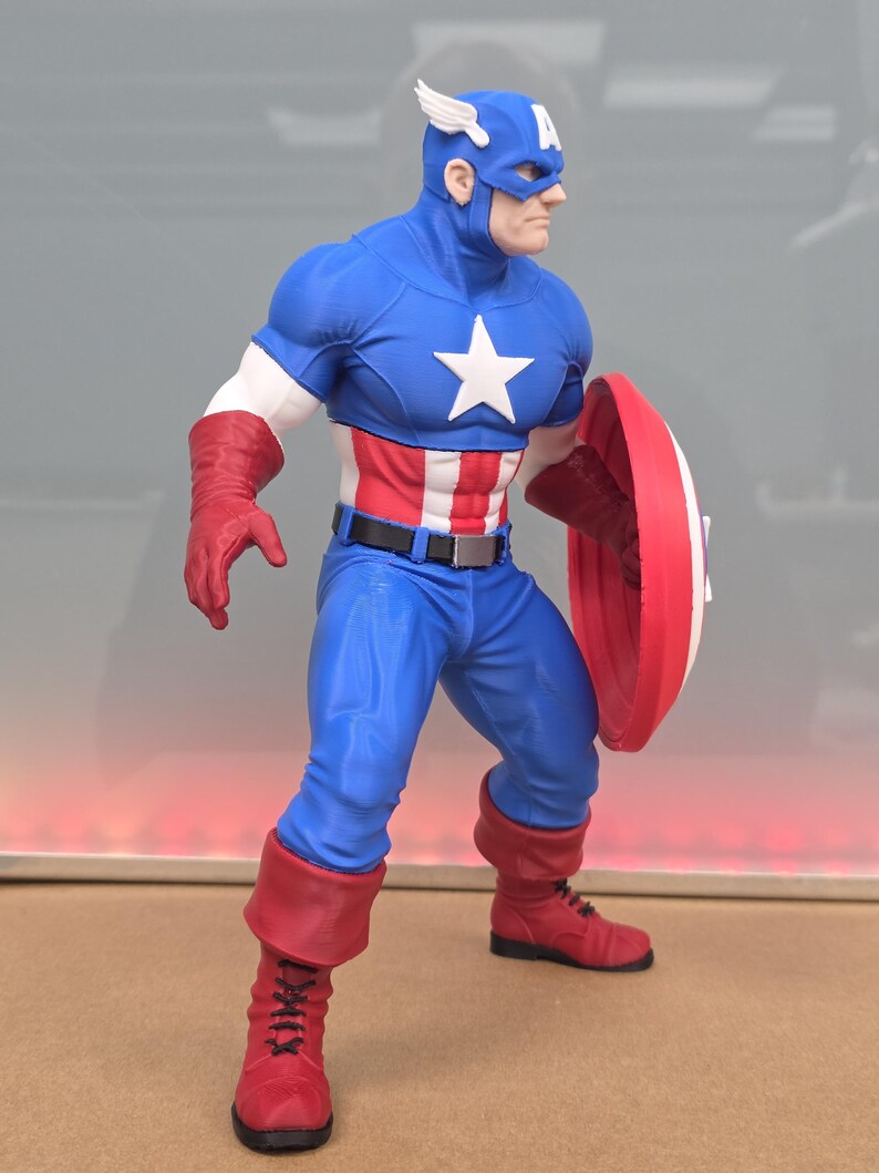 May include: A Captain America action figure wearing a blue suit with a white star on the chest, red and white striped midsection, red gloves, and red boots. The figure wears a blue cowl with a white 'A' and wings, and holds a round red, white, and blue shield.