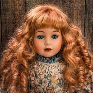 May include: A porcelain doll with long, curly auburn hair and bright blue eyes. The doll wears a floral print dress with a lace collar and a ruffled neckline. The doll's face is painted with delicate features.