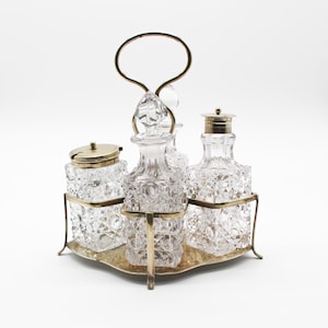 May include: A condiment set featuring three clear, square-cut crystal glass containers with intricate geometric patterns. Two bottles have crystal stoppers, and one jar has a metal lid. The set is held in a brass-toned metal stand with a handle.