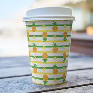 Op de afbeelding: Een papieren wegwerpbeker voor koffie met een witte plastic deksel, voorzien van een herhalend patroon van gele ananassen met groene bladeren in rijen tussen horizontale groene en gele strepen op een witte achtergrond met een fijn ruitpatroon.