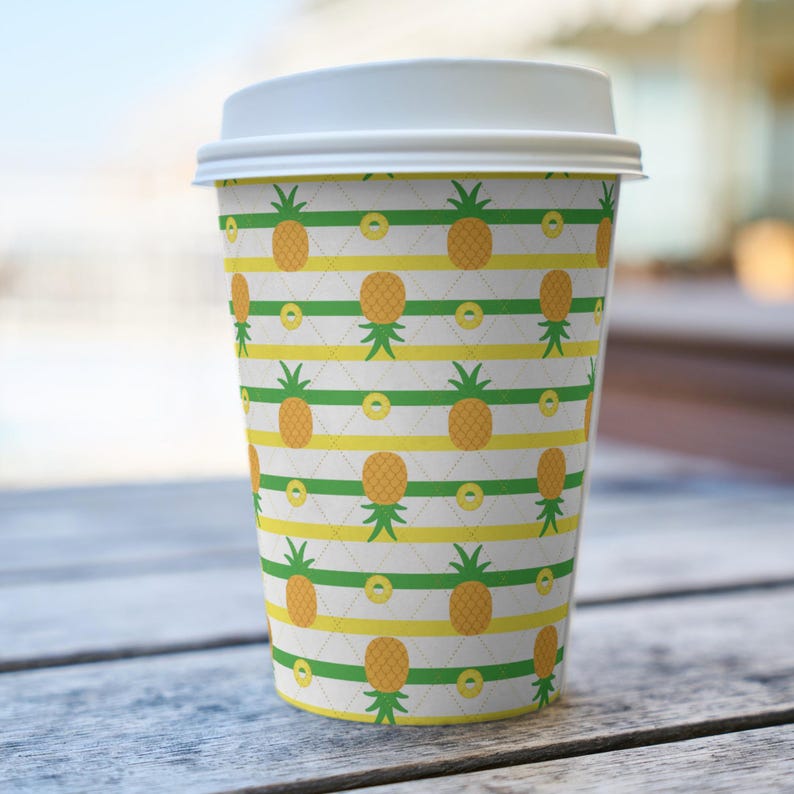 Op de afbeelding: Een papieren wegwerpbeker voor koffie met een witte plastic deksel, voorzien van een herhalend patroon van gele ananassen met groene bladeren in rijen tussen horizontale groene en gele strepen op een witte achtergrond met een fijn ruitpatroon.