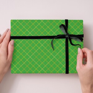Op de afbeelding: Een rechthoekige geschenkdoos verpakt in felgroen papier met een gouden diagonaal ruitpatroon. De doos is gestrikt met een zwart fluwelen lint dat aan de rechterkant tot een strik is geknoopt, wat een contrast vormt met het groen en goud.