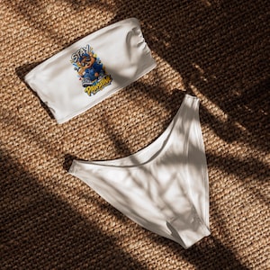 May include: A white two-piece bikini set consisting of a strapless bandeau top and high-cut bottoms. The top features a graphic print of a cartoon bear wearing a blue hoodie and sunglasses with the text STAY PAWSITIVE in yellow and blue.