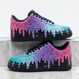 May include: A pair of low-top sneakers featuring a black sole and a gradient upper that transitions from pink to purple to blue. The design includes a black drip pattern overlaying the gradient colors, black laces, and black interior lining.