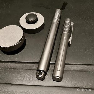 May include: A collection of metallic grey writing implements and accessories. The image shows a pen, a pencil, and two circular objects. The pen and pencil have ridged detailing near the top. The items are on a dark surface.