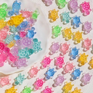 May include: A collection of colorful, translucent turtle-shaped charms in various shades of pink, blue, green, and yellow. The charms are scattered on a white surface, with the text "KEEP HAPPINESS" visible. These decorative charms are ideal for crafting.