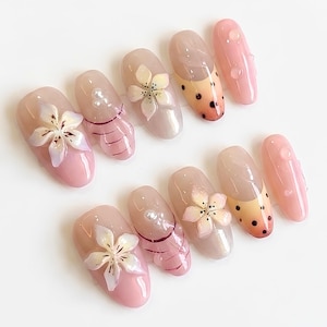 May include: A set of ten press-on nails featuring a glossy finish in shades of soft pink and nude. The designs include three-dimensional white floral appliqués, small white pearls, thin horizontal black lines, and black polka dot patterns.