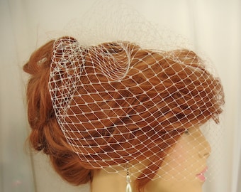 May include: A white birdcage veil with a fine mesh pattern. The veil is worn over a woman's hair, covering her forehead and ears.