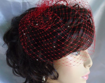 May include: A red birdcage veil with rhinestones, worn over a woman's short, dark hair.