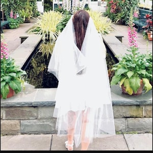 May include: A person wearing a white dress and veil, standing in a garden setting. The veil drapes over the person's head and shoulders. The background features lush greenery, flowers, and a water feature.