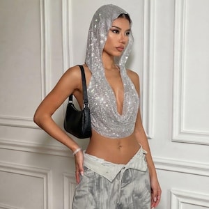 May include: A silver hooded halter top covered in reflective rhinestones with a deep cowl neckline, paired with a black shoulder bag and grey patterned trousers. The top features a metallic mesh-like texture that catches light across the surface.