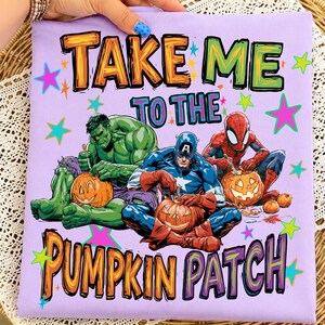 May include: A light purple t-shirt featuring a graphic of the Hulk, Captain America, and Spider-Man carving pumpkins. The text reads "TAKE ME TO THE PUMPKIN PATCH" in orange and purple letters, surrounded by colorful stars on the fabric.