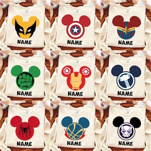 May include: A collection of nine off-white sweatshirts, each featuring a unique graphic of a character-themed Mickey Mouse ear icon. The designs include symbols for Wolverine, Captain America, Captain Marvel, Hulk, Iron Man, Thor, Spider-Man, a basketball, and Black Panther, with the word NAME printed below each.