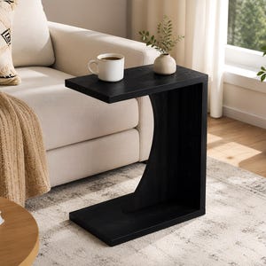 May include: A black C-shaped side table with a flat top and base. A white mug with coffee sits on the table. A beige sofa, patterned pillow, and throw blanket are in the background.