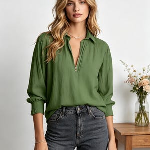 Könnte beinhalten: Olivgrüne Langarmbluse mit Kragen und goldenem Reißverschluss. Die Bluse hat eine lockere Passform und geraffte Manschetten. Das Model trägt dunkelgraue Jeans. Eine Vase mit Blumen steht auf einem Holztisch im Hintergrund.