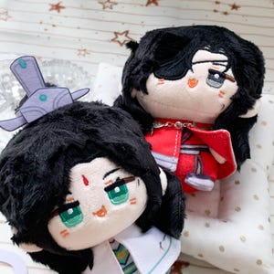 May include: Two plush dolls with black hair and embroidered facial features. One doll wears a purple headpiece and white clothing, while the other has a black eye patch and wears a red outfit with a silver chain detail across the chest.