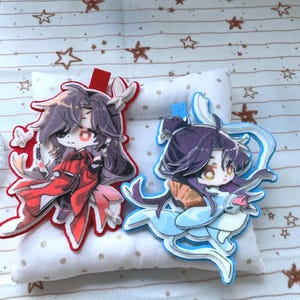 May include: Two layered acrylic charms featuring stylised anime characters. The left charm depicts a character in red robes with dark hair, while the right charm shows a character in light blue robes with dark hair holding a folding fan.