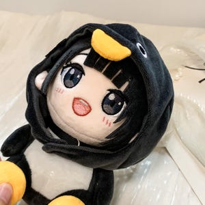 以下が含まれることがあります： 黒髪で大きな目をしたキャラクターのぬいぐるみで、白黒のペンギンをモチーフにしたフード付きの衣装を着ています。フードには黄色いクチバシのディテールがあり、顔は白く、刺繍された目鼻立ちとピンクの頬が特徴です。
