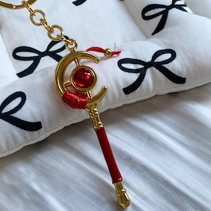 May include: A gold-toned metal keyring featuring a crescent moon shape at the top with a central circular red gemstone. The stem is accented with red thread wrapping and a solid red cylindrical section, ending in a small gold-toned metal tip.
