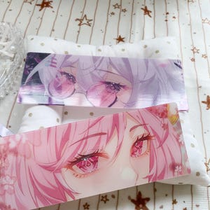May include: Two rectangular acrylic bookmarks featuring anime-style illustrations. The top bookmark displays a character with light purple hair and pink-tinted glasses. The bottom bookmark shows a character with pink hair and detailed pink eyes.
