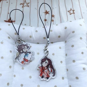 Könnte beinhalten: Zwei durchsichtige Acryl-Anhänger mit Chibi-Anime-Figuren und schwarzen Kordelschlaufen. Der linke Anhänger zeigt eine Figur in weiß-blauem Gewand mit roten Blumen. Der rechte Anhänger zeigt eine Figur in einem rot-orangen Gewand mit langem Haar.