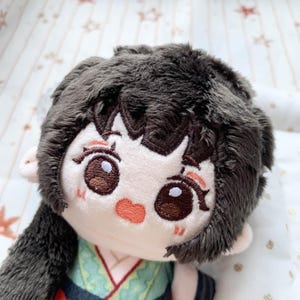 Pode incluir: Uma cabeça de boneco de pelúcia com cabelo felpudo castanho escuro, olhos bordados castanhos com reflexos brancos e um pequeno nariz laranja. O boneco tem uma tez clara e veste uma peça estampada em verde e preto com detalhe de gola vermelha.