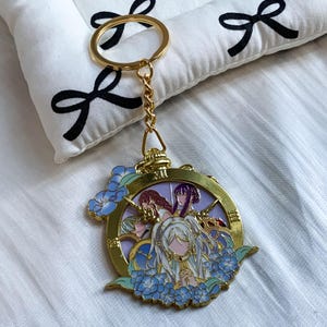 May include: A gold-toned metal keyring featuring a circular frame with Roman numerals. The centre displays an anime-style illustration of three characters with white, purple, and red hair, surrounded by blue floral accents and a light purple background.
