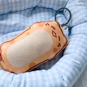 May include: A plush keychain shaped like a rectangular piece of toasted bread with a light brown crust and a tan center. The character features a printed face with closed eyes and a small mouth, attached to a silver metal split ring key holder.