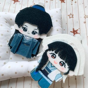 Pode incluir: Dois ornamentos planos de feltro com personagens de estilo anime, olhos grandes e bochechas rosadas. Uma figura tem cabelo preto com faixa azul e túnica azul-escura, a outra tem cabelo preto com tranças e túnica azul-clara.