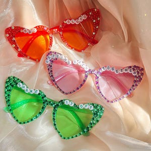 Puede incluir: Cuatro pares de gafas de sol en forma de corazón en rojo, rosa y verde, adornadas con pedrería. Las gafas rojas tienen la palabra "BAD BLOOD". Las gafas rosas dicen "GETTING BACK TOGETHER". Las gafas verdes dicen "SHAKE IT OFF".