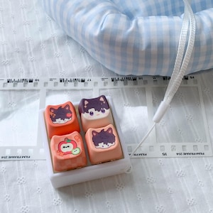 May include: A set of four square mechanical keyboard keycaps housed in a white plastic tray. The keycaps feature cartoon character illustrations with cat ears, including one with an apple motif, in shades of orange, cream, and dark purple.