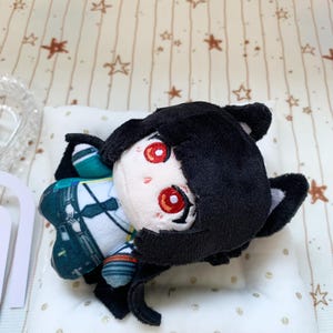 May include: A plush doll featuring a character with short black hair, cat ears, and bright red eyes. The doll wears a white shirt with a black tie, a dark green vest, and dark trousers. The plush has a soft, rounded face and is positioned on a white pillow.