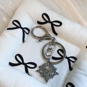 May include: A silver-toned metal keychain featuring a circular split ring and a lobster claw clasp. Attached to the ring is a star-shaped charm with an intricate, cut-out geometric pattern in the center, showcasing a detailed, symmetrical design.
