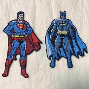 Puede incluir: Parches bordados de Superman y Batman. Superman viste un traje azul con capa roja y el logo "S". Batman lleva un traje azul y negro con un símbolo de murciélago y una larga capa. Ambos parches tienen bordes negros.