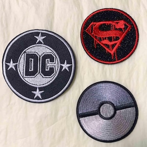 Może przedstawiać: Trzy okrągłe, haftowane naszywki. Jedna przedstawia logo DC Comics w czerni i bieli, druga logo Supermana w kolorze czerwonym i czarnym, a trzecia szaro-czarny wzór Pokeballa. Idealne do personalizacji odzieży lub akcesoriów.