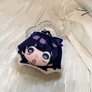 May include: A plush keychain featuring a stylized anime character head with dark blue hair, large round eyes, and an open mouth expression. The character has a pale complexion and small red accents in the hair. A black cord loop is attached to the top.