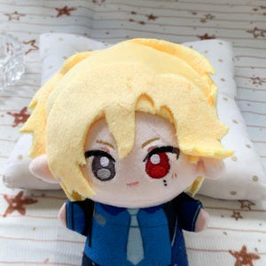 May include: A plush doll featuring bright yellow hair, one grey eye, and one red eye. The doll wears a dark blue collared shirt with a light blue tie. A small black mark is visible under the red eye, and the doll has a soft, rounded face.