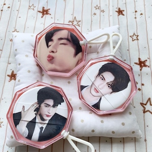 May include: Three octagonal pink plastic handheld mirrors, each featuring a different photographic portrait of a person with dark hair. The mirrors have a white loop handle at the top or side and a clear frame surrounding the circular image insert.