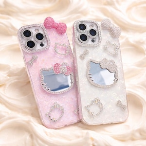 May include: Two bedazzled phone cases. One is pink with a pink bow and a Hello Kitty mirror. The other is white with a silver bow and a Hello Kitty mirror. Both cases have silver rhinestone accents.