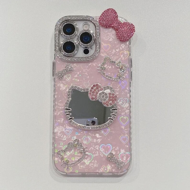 May include: Pink phone case with a sparkly bow and Hello Kitty designs. The case features a mirror and is adorned with rhinestones around the camera and Hello Kitty details. The background has a heart pattern.