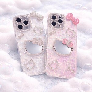 May include: Two phone cases with a Hello Kitty design. One case is white, the other is pink. Both cases have a mirrored Hello Kitty face, bow, and other decorative rhinestones. The cases are surrounded by bubbles.