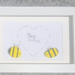 May include: A white framed print with a bee-themed design. The artwork shows two cartoon bees with yellow and black stripes, and the words "Bee mine" inside a heart outline. The frame is white.