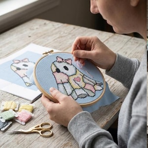 Op de afbeelding: Een ronde houten borduurring met lichtblauwe stof en een kruissteekontwerp van een wit wezen met een roze strik, gele accenten en een klein roze hartje. Een naald met roze draad is in de stof gestoken.