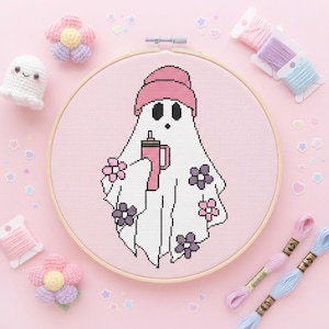May include: A cross-stitch embroidery in a round wooden hoop featuring a white ghost wearing a pink beanie and holding a pink tumbler. The ghost is decorated with small pink and purple flowers on a light pink fabric background, surrounded by yarn.