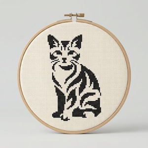 以下が含まれることがあります： 白いアイーダ布に黒い猫のシルエットをクロスステッチで刺繍し、円形の木製刺繍枠に固定したもの。猫のデザインはピクセルアート風に表現されており、耳の尖った猫が座っている様子が描かれています。