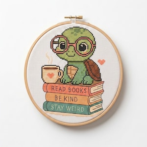May include: A circular wooden embroidery hoop containing a cross-stitch pattern of a green turtle wearing pink glasses. The turtle sits on a stack of three books labeled READ BOOKS, BE KIND, and STAY WEIRD, holding a mug with a small heart icon.