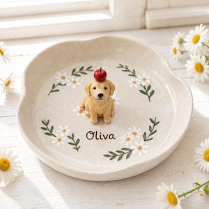 Peut inclure: Un vide-poche rond en céramique blanc cassé à bord festonné, orné au centre d'une figurine de golden retriever peinte à la main avec une petite pomme rouge sur la tête. Le plat est décoré de fleurs de marguerites blanches et du nom Olivia.